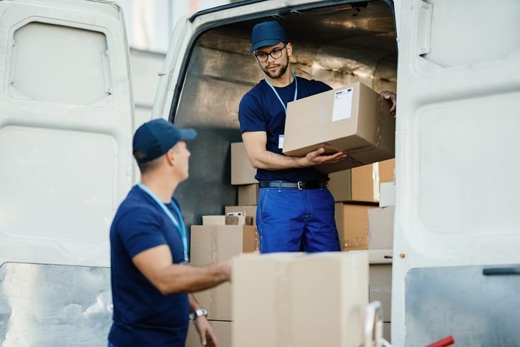 Young worker loading cardboard boxes in a delivery van and communicating with his colleague.